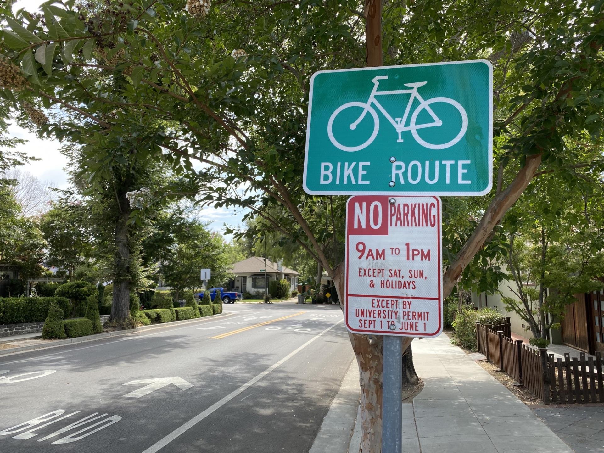 A bike route sign and a parking sign that reads, "No parking. 9 a.m. to 1 p.m., except Saturday, Sunday, and holidays. Except by university permit. September 1 to June 1."