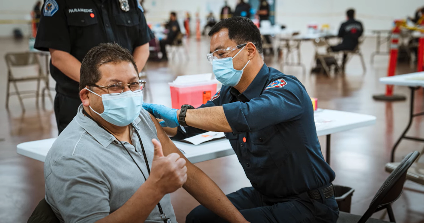 A man wearing a mask getting a COVID-19 shot in the left arm by a San Jose Fire Department firefighter