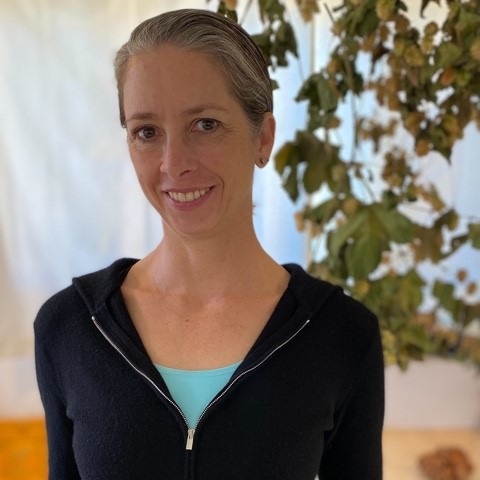 Woman with light complexion and blonde-grey hair pulled back. She is wearing a black zipper pullover with a turquoise shirt underneath. She is also wearing studded earrings.