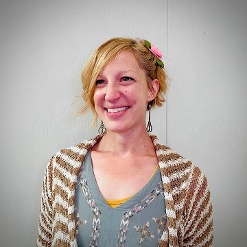 Woman with a light complexion and short blonde hair with a pink flower barrette on the right side of her head. She is wearing a brown and white striped knitted cardigan and a blue-grey-neck short with embroidered patterns on it. She is also wearing scissor shaped earrings.