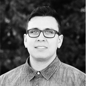 Black and white photo of a man with short dark hair wearing glasses and a denim collared shirt with a white button.