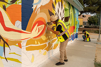 A multicolored mural being painted by volunteers wearing yellow vests