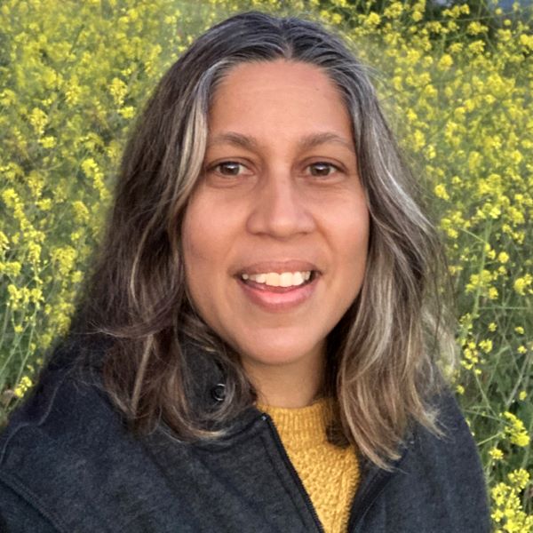 Image of a woman with black and grey hair shoulder length. She is in front of a yellow floral bush. She is wearing a dark fleece and yellow sweater.