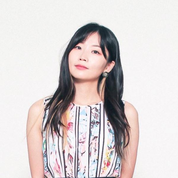 Woman with dark hair below the shoulders with head tilted. Wearing a sleaveless blouse with a floral pattern. Her hands are behind her back. The background is a light white color.