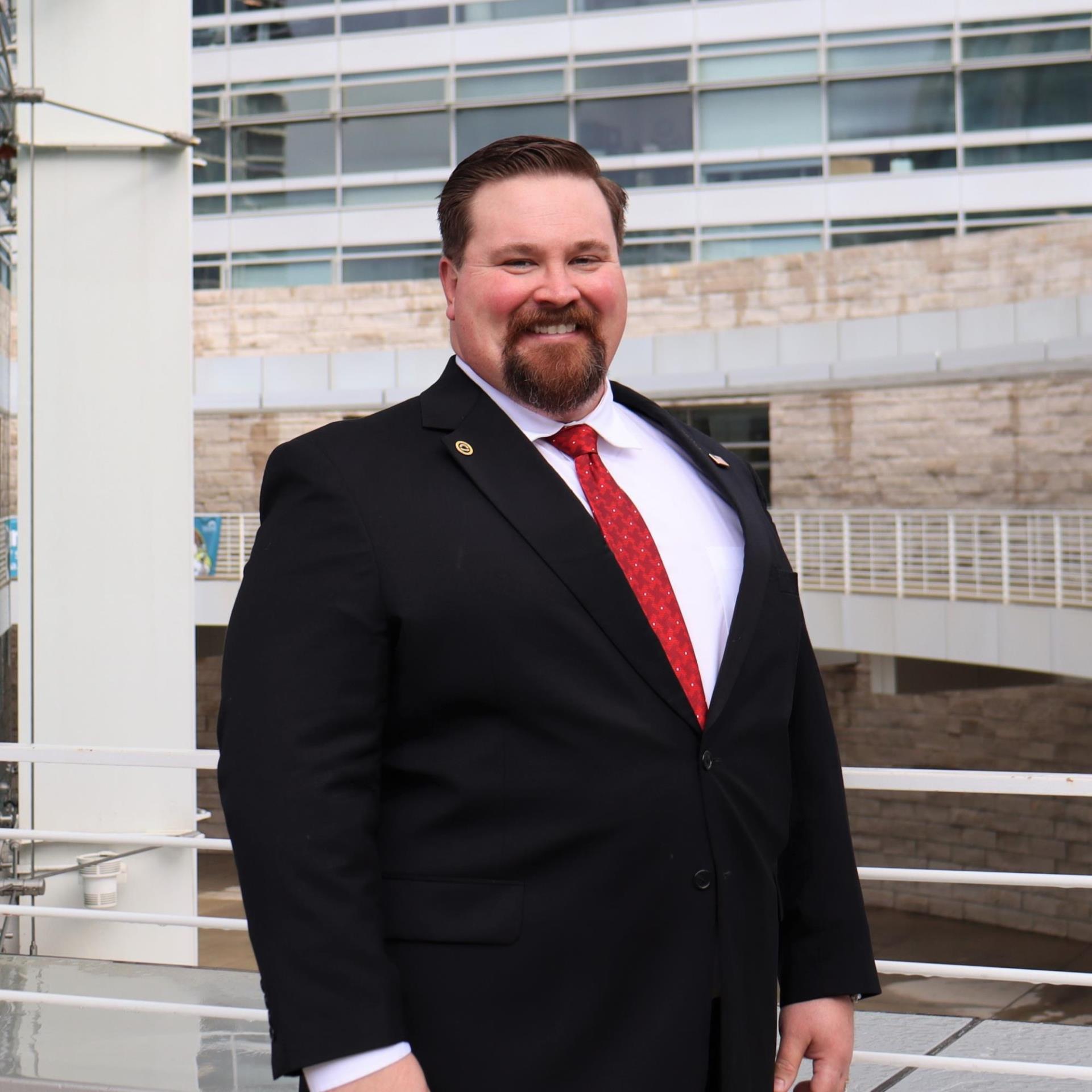 Picture of Jonathan Fleming wearing a black blazer, a white button up, and a red tie, smiling in front of San Jose City Hall.