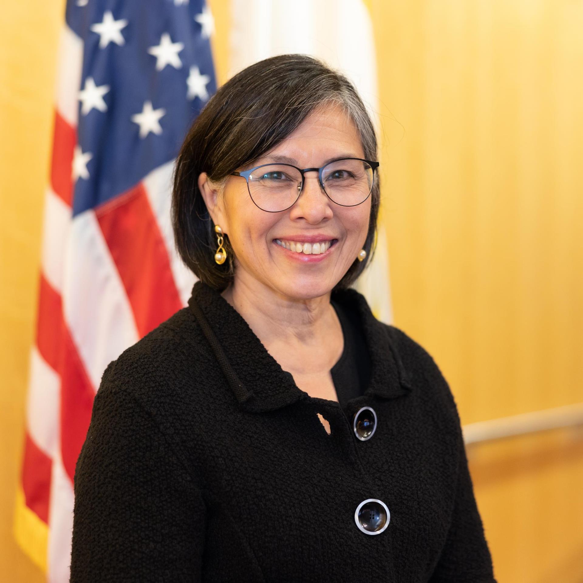 Rosemary Kamei, a woman smiling with medium black hair and glasses wearing a black coat posing in front of a US flag inside San Jose City Council chambers.