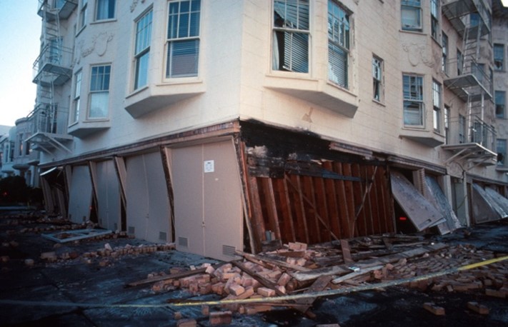 Photograph of multistory building with the first floor partially collapsed and building debris spilling onto sidewalk and street.