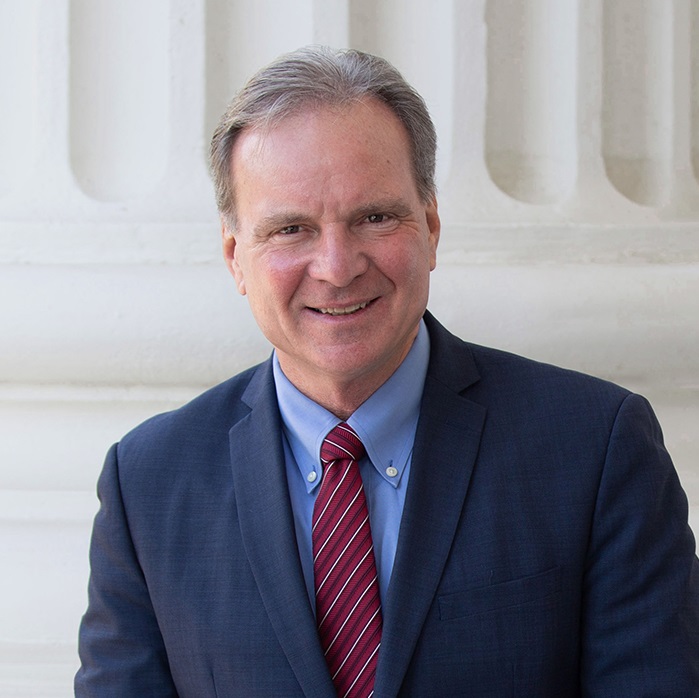 Dave Cortese, a man smiling with greyish-brown hair wearing a blue suit, blue shirt, and red tie in front of the California State Capitol building.