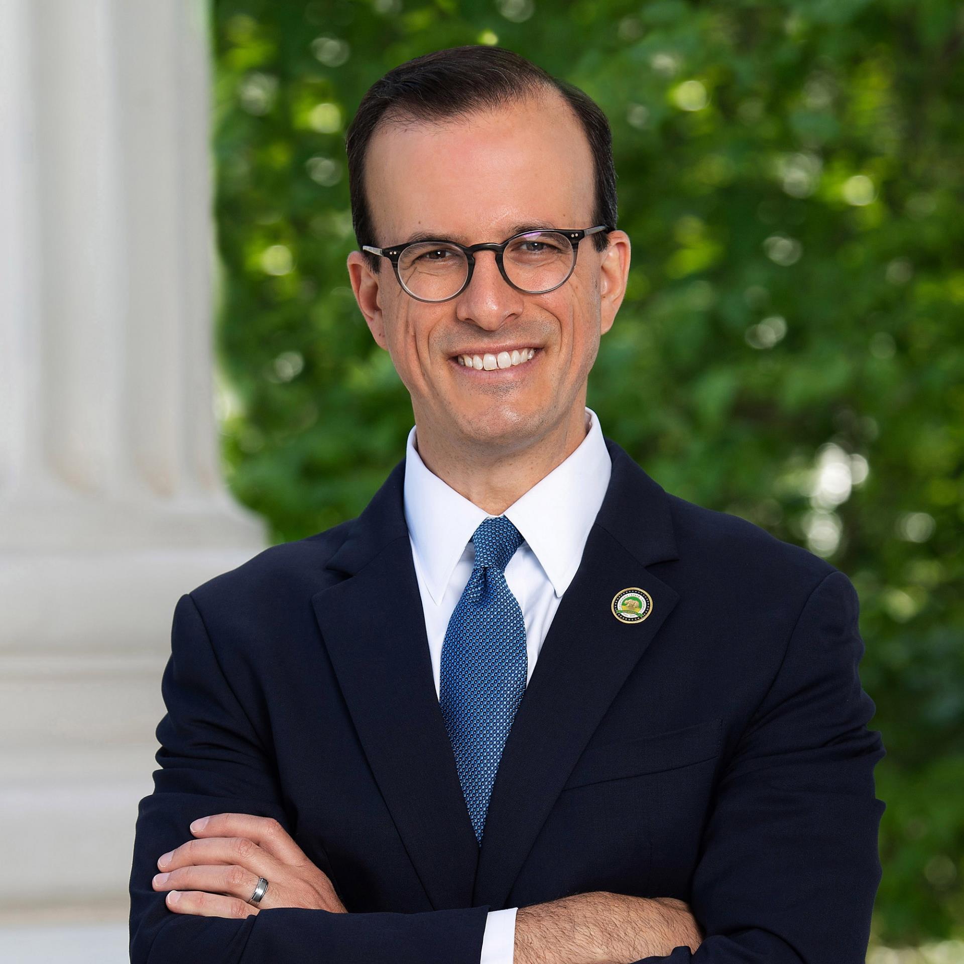 Marc Berman, a man smiling with arms crossed wearing glasses, with short brown hair, wearing a dark navy suit, white shirt and blue tie, posing in front of the California State Capitol and trees.