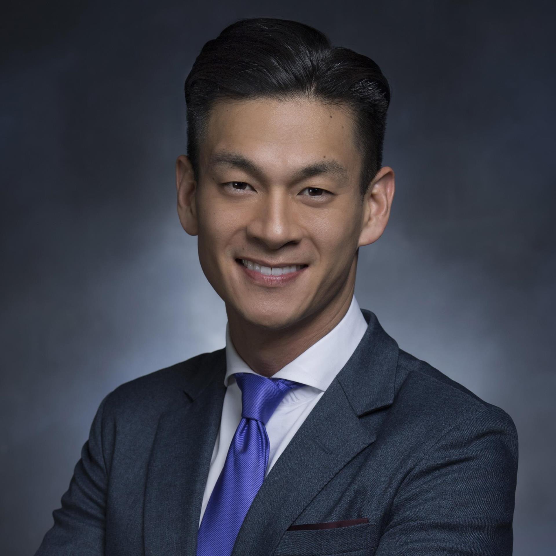 Evan Low, a man smiling with short black hair wearing a dark navy suit, white shirt, and blue tie posing in front of a plain grey yearbook-style background.