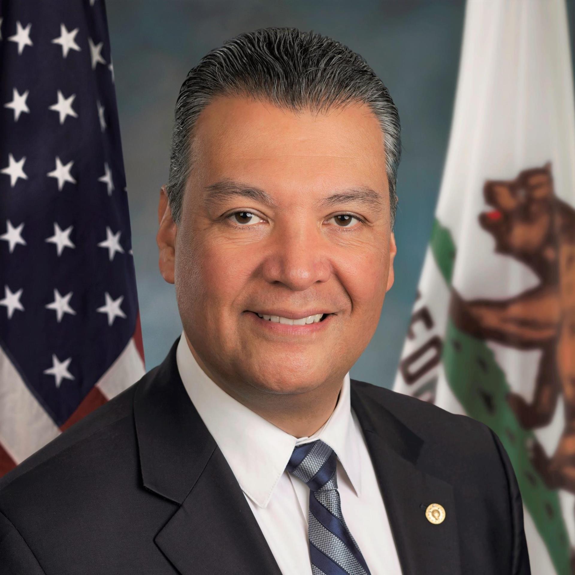 Alex Padilla, a man smiling with salt and pepper hair, a grey suit, white shirt and dark tie posing in front of the US and California flags.