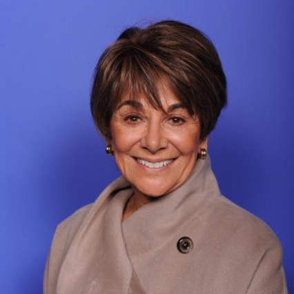 Anna Eshoo, a woman smiling with short brown hair wearing a tan coat posing in front of a plain blue background.