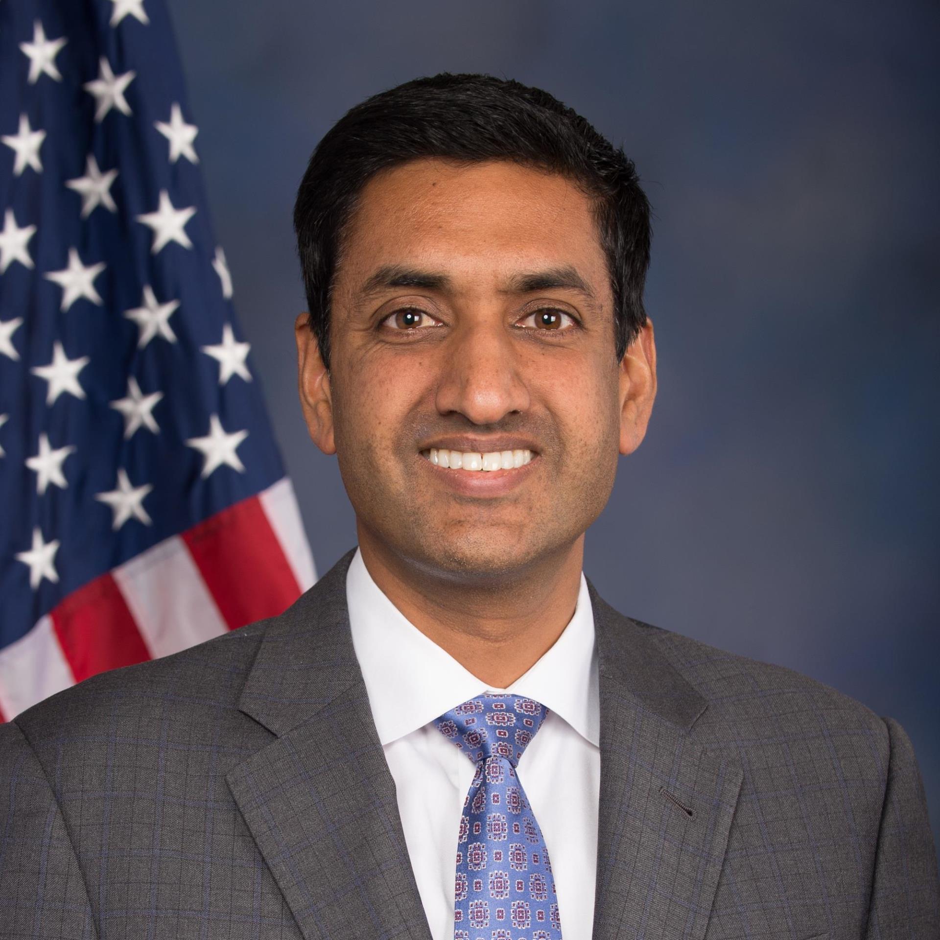 Ro Khanna, a man smiling with short black hair wearing a grey suit, white shirt, and blue tie posing in front of the US flag.