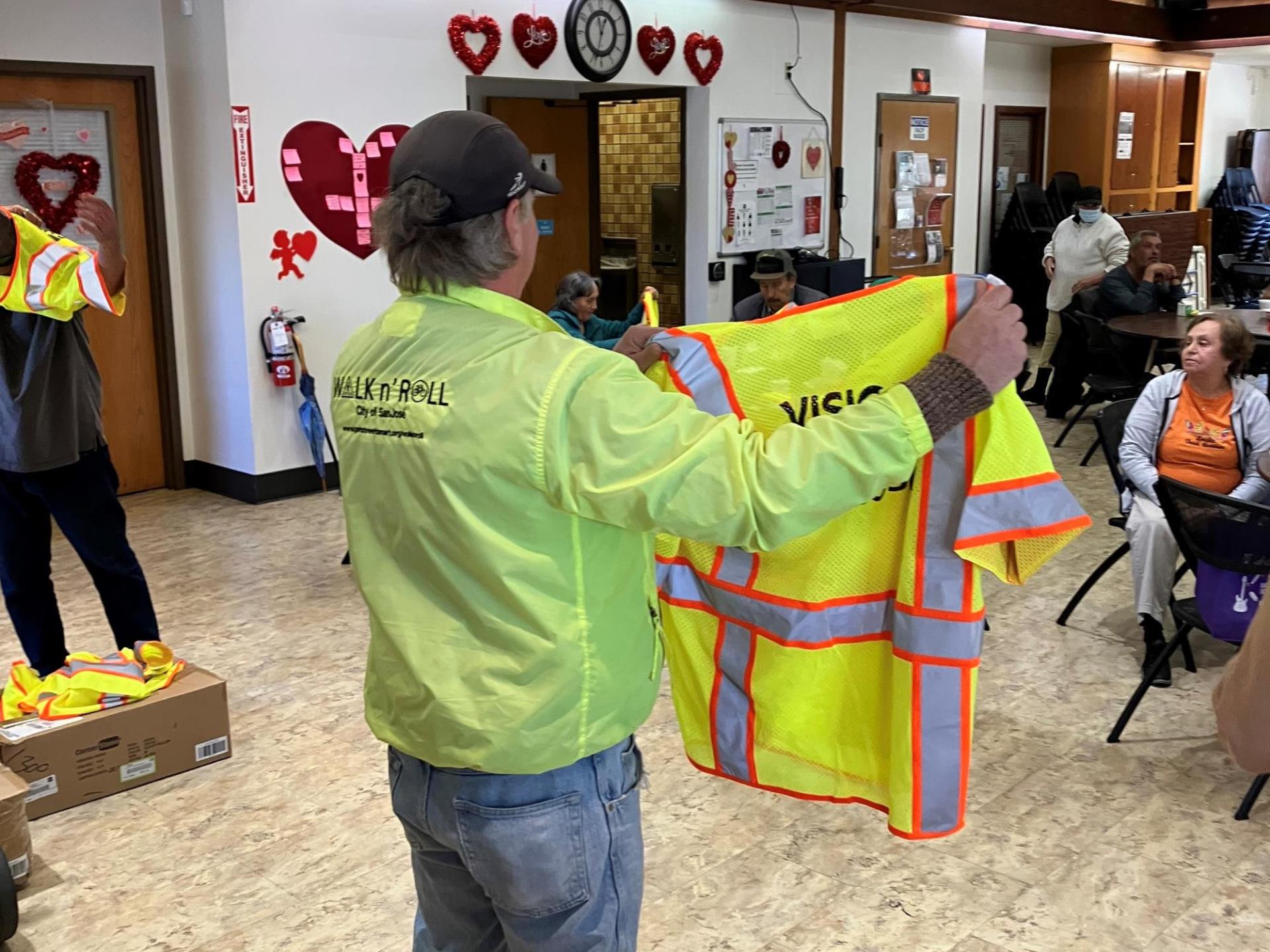 A man in a bright yellow jacket holds up a bright yellow vest in front of a crowd at a community center