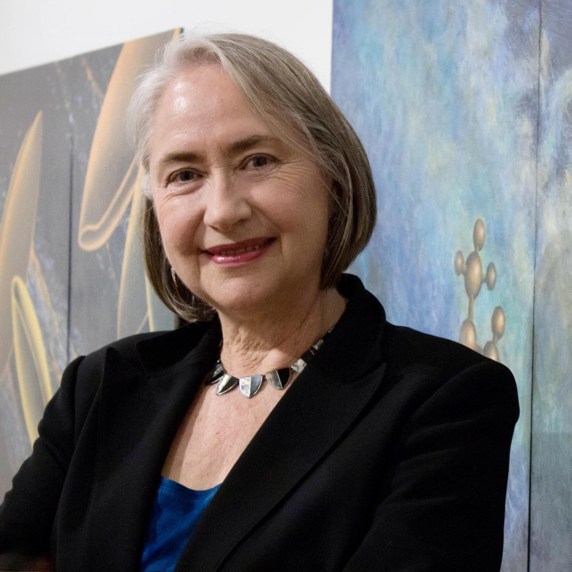 Photo of a white woman with grey hair smiling with a painting behind her.