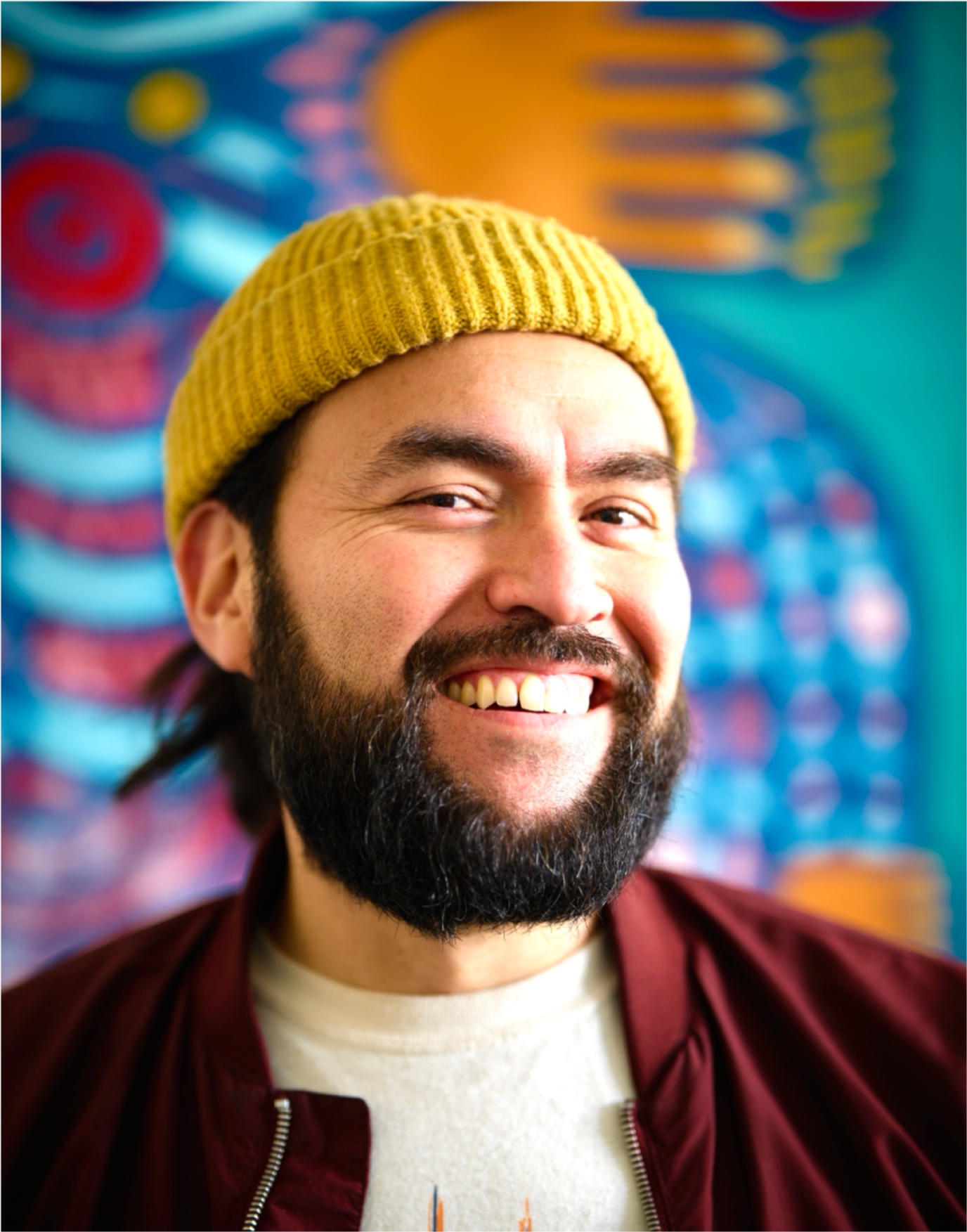 Portait of a latino male in his mid-30s with a beard ad mustache wearing a yellow hat in front of a painting.