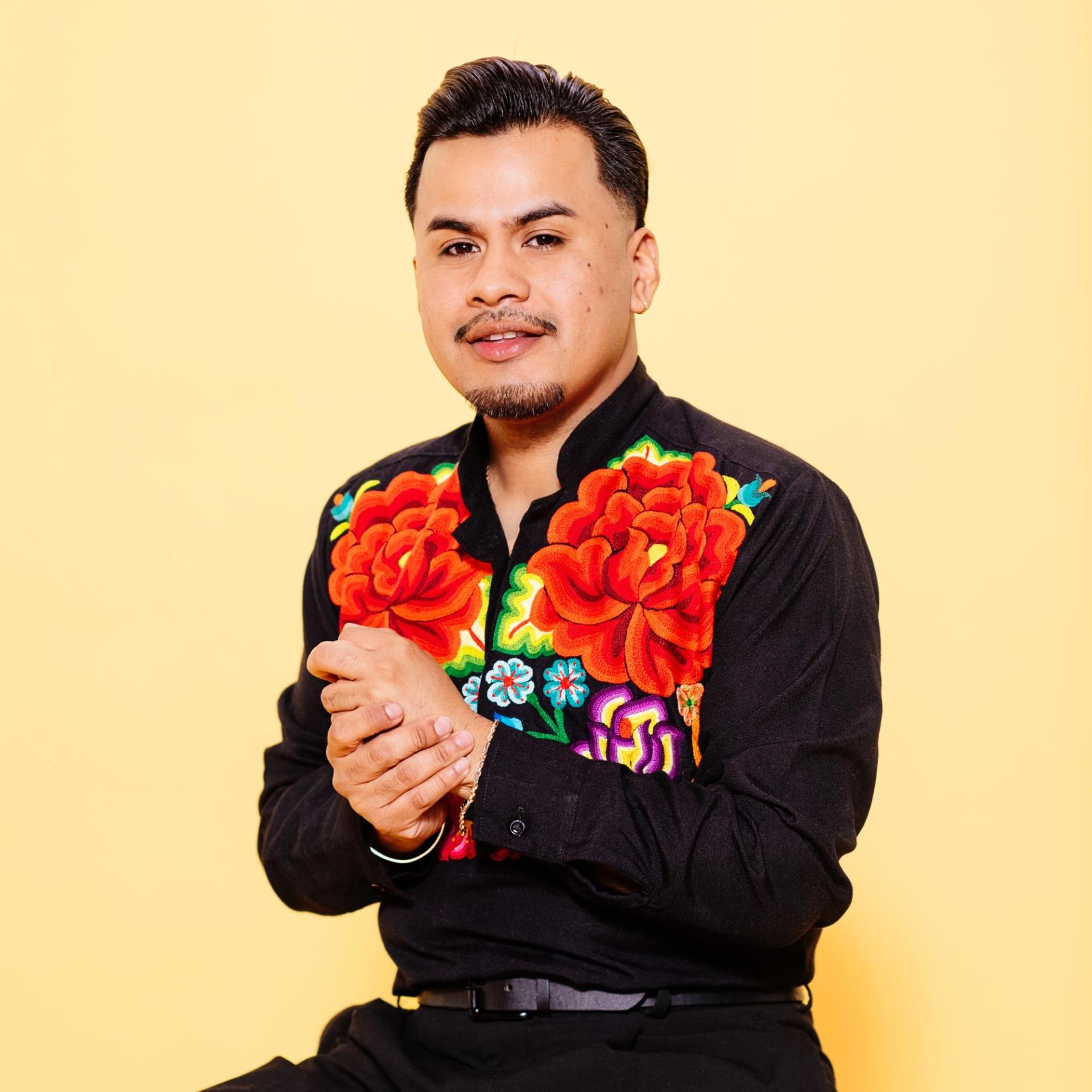 Headshot of a Latino man with a thin mustache wearing a black shirt with roses on it.