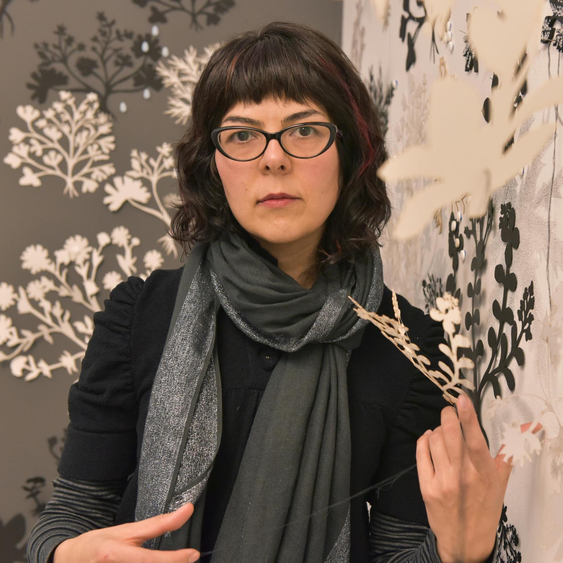 Photo of a middle-aged woman with dark hair and glasses wearing a grey scarf. She is in front of a botanical grey and white print. She is holding a botanical item in her right hand.