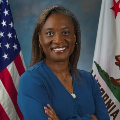 Laphonza Butler, a woman smiling with arms crossed with long brown hair wearing a blue dress posing in front of the US and California flags.