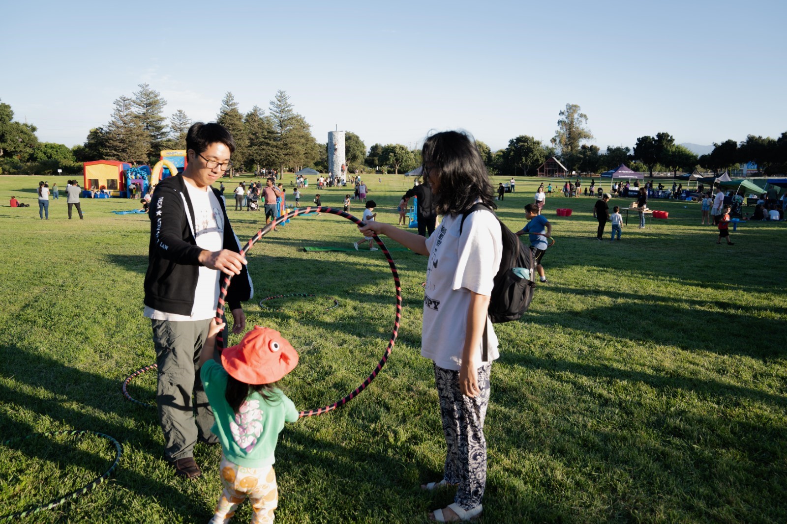 Two adults and one child play together with a hula hoop on a park lawn. Behind them are several more families playing on the lawn, pop-up tents, and a bouncy house