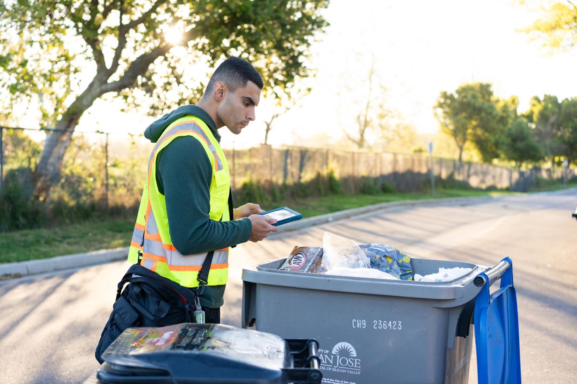 A man in a green hoodie with a neon safety vest over it is holding an iPad while inspecting the contnets of an open recycling bin.