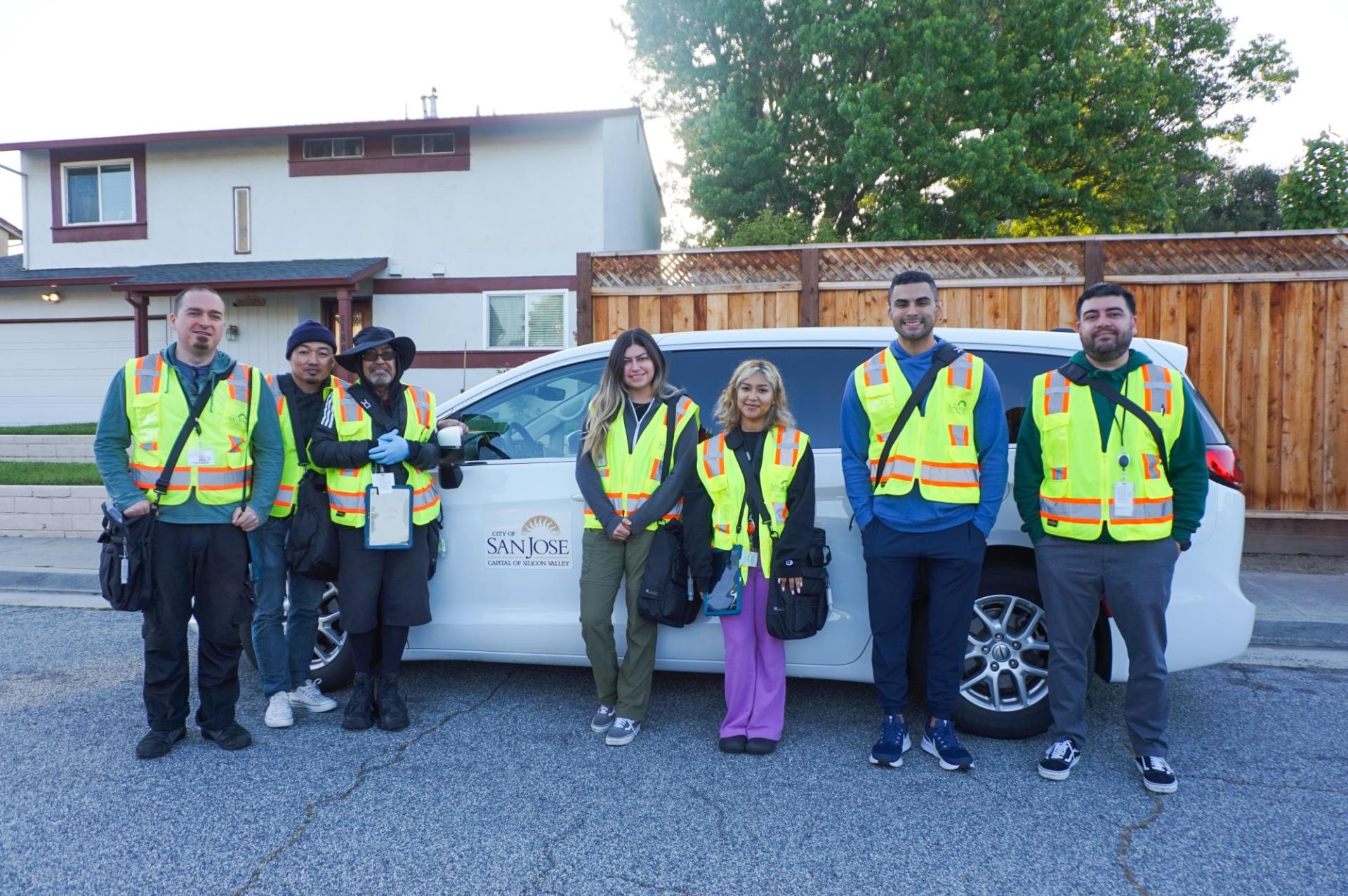 Seven City of San Jose employees are standing in front of a white mini van with the City of San Jose's logo on it. The employees are wearing neon safety vests over their clothes and have a bag strapped over their chest. One employee is wearing plastic gloves.