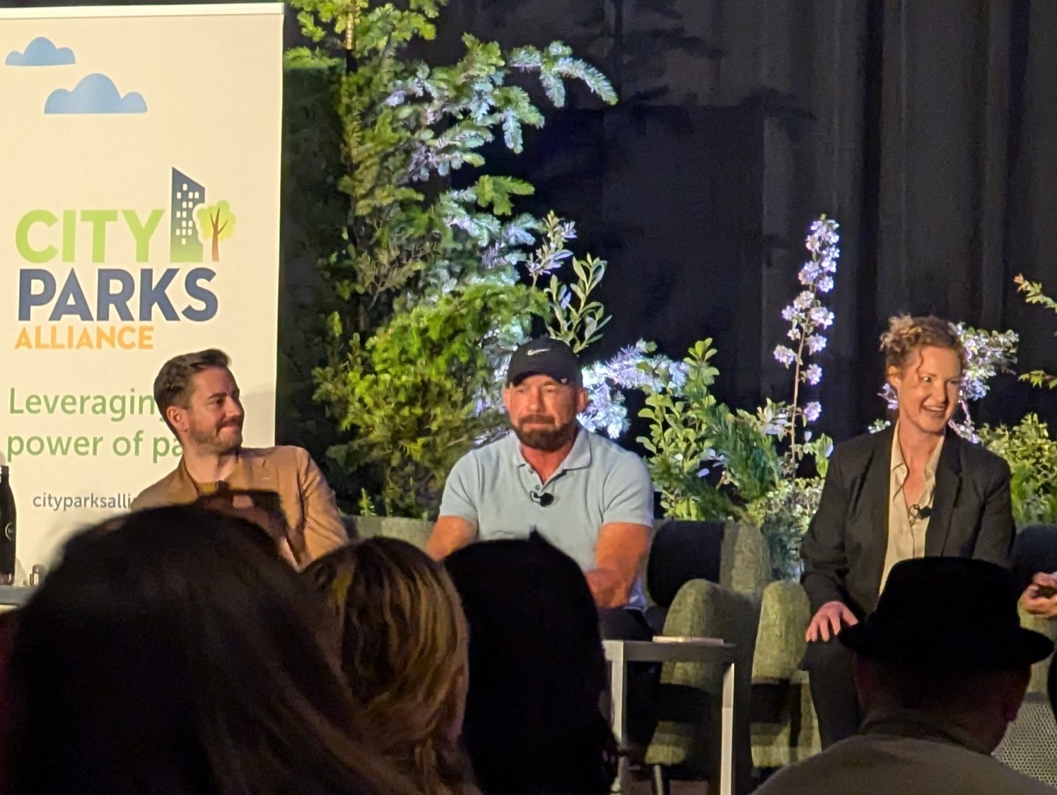 Three speakers are seated in front of an audience at the Greater and Greener Conference. Left to right, they are a conference facilitator, Paul Pereria, and Liz Sewell.