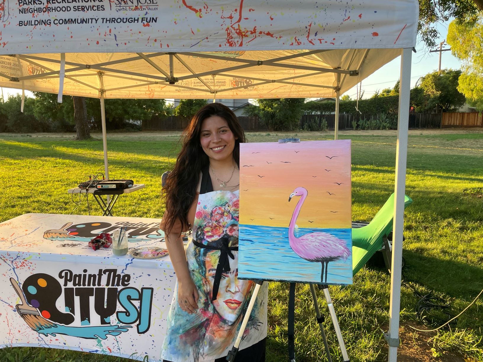 San José Artist and Instructor, Luz Gonzalez, smiles as she stands with her completed canvas at Noble Park. The painting is a pink flamingo with a colorful sunset and ocean background.