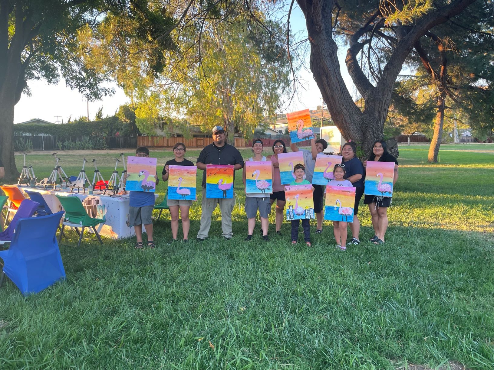 Ten youth and adult participants pose with their completed canvases at Noble Park. The paintings are of a pink flamingo with colorful sunset and ocean backgrounds.