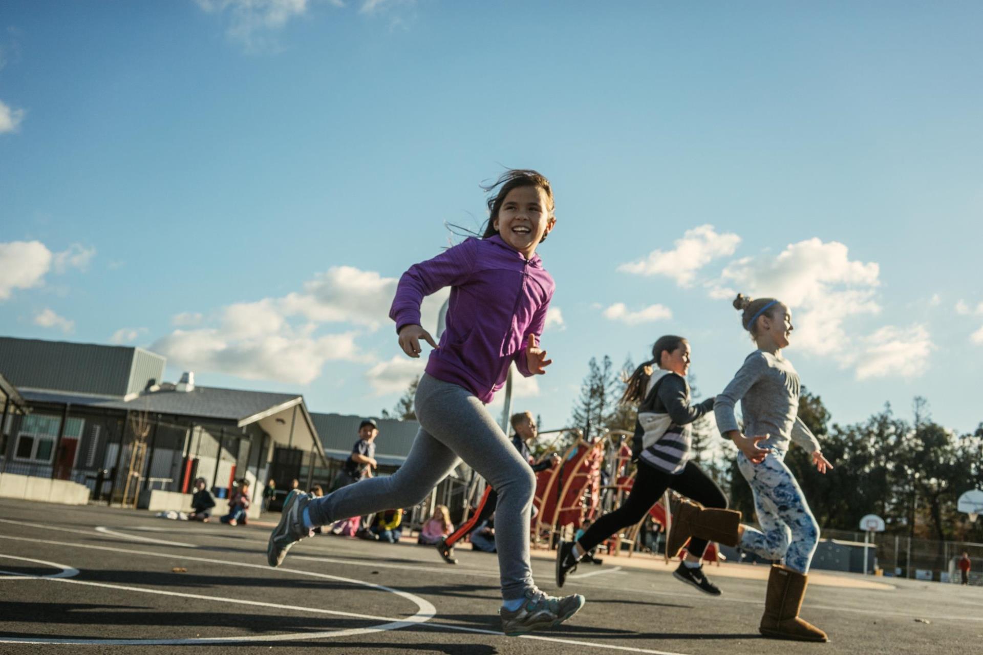 A group of smiling girls run across an elementary school's blacktop. In the background, other children play on a climbing structure