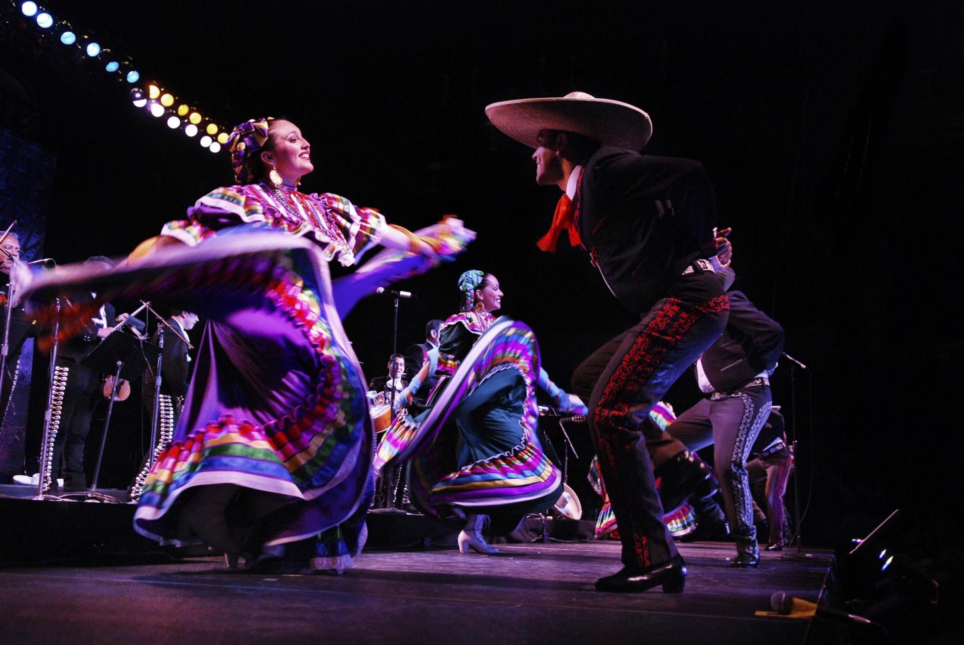 One woman smiling while dancing and twirling a colorful dress. Facing the woman is a man in a large sombrero smiling wearing black and white.  