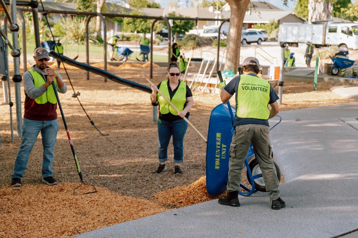 Three volunteers wearing yellow hi-vis vests stand in a playground area. Two hold rakes, one is using a blue wheelbarrow to dump wood chips